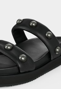 Μαύρο σανδάλι slide με δύο πλατιά επενδεδυμένα λουριά που διαθέτουν μεγάλα στρογγυλά μεταλλικά καρφιά και υφή στην σόλα.