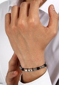 Svart och silverarmband med metallaccenter, cylindriska former och texturerade pärlor. Buren på en hand mot en vit skjorta.