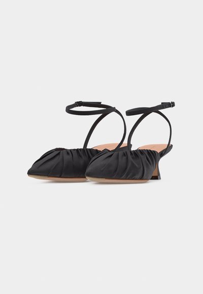 Μαύρες γόβες slingback με ραμμένο ύφασμα, μυτερές, με λεπτές λουρίδες στον αστράγαλο και χαμηλό χοντρό τακούνι σε απλό φωτεινό φόντο.