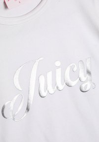 Biela bavlnená tričko s veľkým kovovým strieborným kurzívnym logom "Juicy" na prednej strane, s klasickým okrúhlym výstrihom a hladkou textúrou.