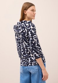 Sudadera con capucha de estampado de leopardo en tonos azul marino y blanco, con mangas de tres cuartos, textura de tela suave y corte holgado. Combinada con jeans azul claro.