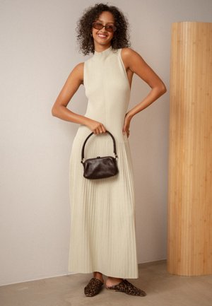 Kobieta o kręconych włosach w okularach przeciwsłonecznych, w kremowej plisowanej sukience bez rękawów, w butach w panterkę, trzymająca małą brązową torebkę, uśmiechająca się w pomieszczeniu.