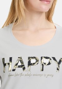 Grijze t-shirt met het woord "HAPPY" in patroon zwarte en groene letters, versierd met zilveren nagels en een gouden tekst citaat eronder.