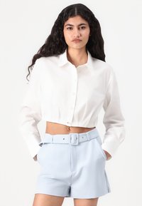 Camicetta bianca a maniche lunghe, corta e con bottoni, abbinata a pantaloni corti a vita alta di un blu chiaro con cintura e tasche laterali.