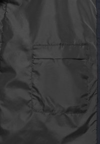 Schwarzer Stoff mit einer rechteckigen Aufsatztasche, die oben einen horizontalen Klappenverschluss hat.