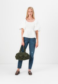 Blusa bianca a texture con maniche a palloncino, abbinata a jeans blu e una borsa in corduroy verde con due tasche frontali e dettagli in oro.