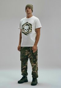 Biała koszulka z grafiką w hexagonalny wzór kamuflażu z czaszką; połączona z zielonymi spodniami cargo w kamuflażu i czarnymi butami sportowymi.