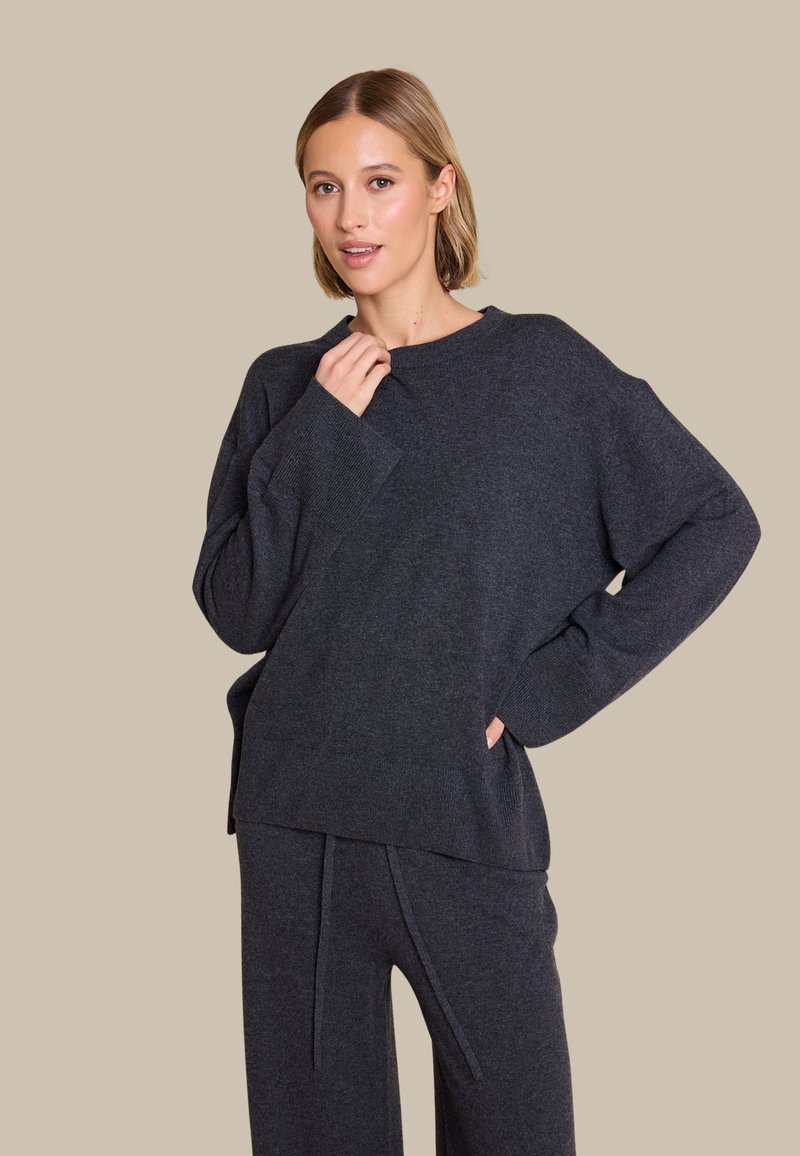 Mørk grå chunky strikkegenser med avslappet passform, brede ermer og ribbestikking ved håndleddene, kombinert med matchende bukser i lignende farge.