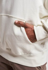 Lys beige hettegenser med frontlomme. Har ribbestrikkede mansjetter og kant. Teksturert stoff, synlig søm langs lommeåpningen.
