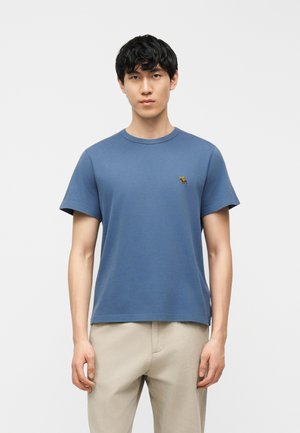 Młody mężczyzna ubrany w niebieski t-shirt z krótkim rękawem z małym logo łosia na piersi i beżowe spodnie, stojący na jednolitym tle.