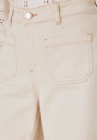 Pantalon beige clair taille haute en tissu robuste, avec surpiqûres orange contrastantes, deux poches avant et une fermeture à bouton métallique.