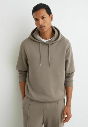 Man die een taupekleurige hoodie en bijpassende broek draagt, staand met handen in de zakken tegen een effen witte muur.