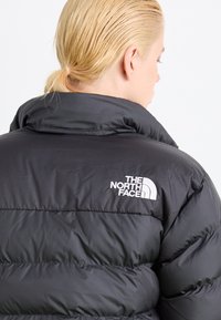 Schwarze Daunenjacke mit horizontalem Steppmuster, ausgestattet mit hohem Kragen und weißem Logo auf der linken Schulter. Glatte, leichte Textur.