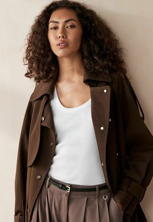 Brun trenchcoat med en strukturerad design, med stora knappar, lagerkrage och en mjuk textur över en vit linne och veckade byxor.
