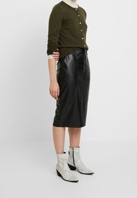 Cardigan verde oliva con chiusura a bottone, abbinato a una gonna a matita in pelle nera e stivaletti alla caviglia bianchi con stampa serpente.