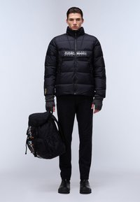 Schwarze Pufferjacke mit hohem Kragen, mit einem Front-Logo-Patch. Schwarze Hose und fingerlose Handschuhe. Trägt einen schwarzen Rucksack mit bunten Akzenten.