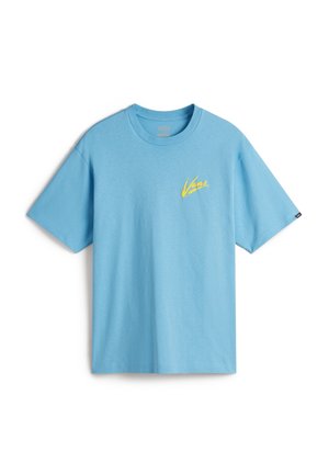 Persoon draagt een lichtblauw Vans T-shirt met korte mouwen over een wit T-shirt met lange mouwen, met donkerbruine broek, handen in de zakken.