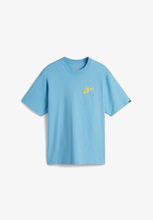 Persoon draagt een lichtblauw Vans T-shirt met korte mouwen over een wit T-shirt met lange mouwen, met donkerbruine broek, handen in de zakken.