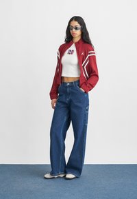 Czerwona sportowa bomberka z białymi paskami, biała krótka bluzka na ramiączkach z napisem "23", szerokie niebieskie dżinsy oraz jasne trampki.
