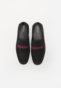 Sorte slip-on sko med glatt tekstur, bred stropp med blå og røde striper, og design med rund tå.