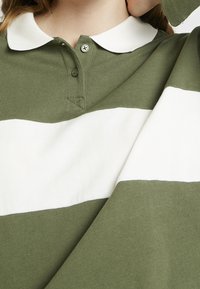 Personne portant un polo à manches longues rayé vert et blanc avec un col blanc, main levée près du visage, gros plan sur le buste.