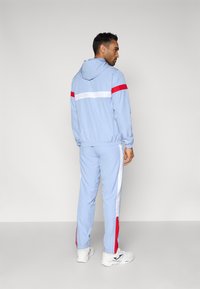 Ανοιχτό μπλε φόρμα με κόκκινες και λευκές ρίγες. Το μπουφάν έχει κουκούλα, ελαστικά μανίκια και μέση. Ορατή λεπτομέρεια με λογότυπο στο πίσω μέρος.