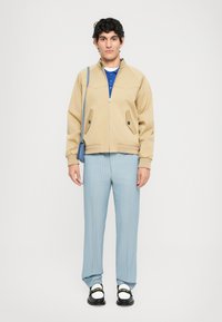 Mlad moški stoji oblečen v bež jakno na zadrgo, modro polo majico, svetlo modre hlače z zebraste črte, črne loafer čevlje z belimi nogavicami in modro torbo preko ramena.