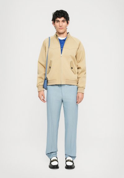 Young man standing wearing beige zip jacket, blue polo shirt, light blue pinstripe pants, black loafers with white socks, and blue shoulder bag.