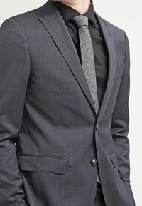 Giacca da completo grigio scuro con vestibilità slim, rever a punta e due tasche frontali, abbinata a una cravatta grigia strutturata sopra una camicia nera.