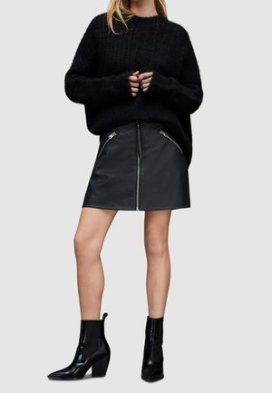 Femme portant un pull noir duveteux, une mini-jupe en cuir noire avec une fermeture éclair à l'avant et des poches zippées, ainsi que des bottines noires à talons.