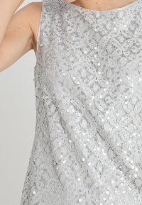 Haut en dentelle gris avec un motif floral, agrémenté de sequins, présentant un col rond et un design sans manches. Fini texturé.