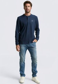 Tumsas zils henleja krekls ar pogu joslu, kombinēts ar gaiši zilām džinsa biksēm un baltām sporta kurpēm, ko valkā stāvošs vīriešu modelis.