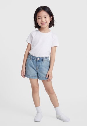 Uśmiechnięta dziewczyna o krótkich czarnych włosach, ubrana w białą koszulkę, szorty dżinsowe, białe skarpetki i białe buty wsuwane, stojąca na jednolitym tle.