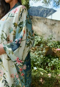 A woman stands outdoors partially obscured by a floral garment, surrounded by greenery and vibrant flowers under a clear sky.