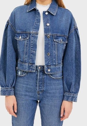 Vrouw draagt een blauwe spijkerjas met knoopsluiting over een wit geribd shirt, gecombineerd met hooggetailleerde blauwe spijkerbroeken.