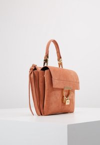 Różowa torebka z zamszu o wyraźnie zdefiniowanej sylwetce, złotymi elementami, zapięciem na klapę oraz charakterystycznym metalowym akcentem z przodu.