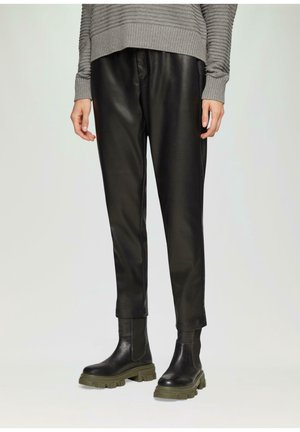 Osoba nosící černé kožené kalhoty, šedý žebrovaný svetr a černé masivní boty se zelenými podrážkami, stojící před jednobarevným pozadím.