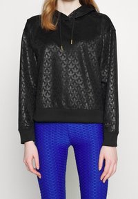 Sudadera negra con capucha y un patrón de hojas texturizadas, con cordones y puños acanalados. Combinada con leggings azules texturizados.