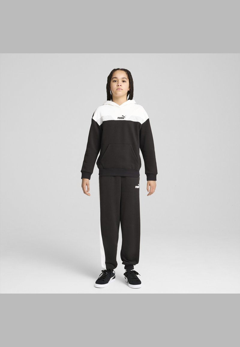 Chándal PUMA en blanco y negro que incluye una sudadera con capucha y un bolsillo canguro, así como pantalones jogger con franjas laterales blancas. Zapatillas negras.