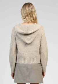 Pull à capuche beige avec une bordure côtelée, présentant de subtils embellissements aux manches et une texture en tricot douce. Porté par-dessus une jupe grise.
