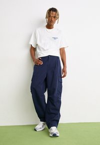 Tricou alb cu logo-ul albastru "Carhartt WIP", asortat cu pantaloni cargo bleumarin cu buzunare laterale, purtați cu adidași albi.
