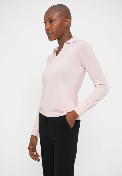 Lichtroze longsleeve polo met een V-hals ontwerp. Zachte, gladde textuur, getailleerde silhouette, gecombineerd met zwarte broek, handen in de zakken.