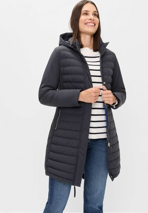 Woman wearing a black quilted hooded coat over a white and black striped sweater and blue jeans, smiling and holding the coat's zipper.