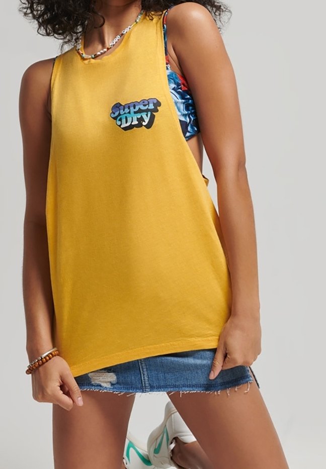 Top giallo senza maniche con il logo "Super Dry", abbinato a una gonna di jeans strappata. Il modello indossa un reggiseno a fantasia mista sotto. Scarpe sportive casual.