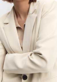 Blazer entallado beige con una textura suave, que presenta un solo botón negro, solapas anchas y mangas estilizadas, superpuesto a un top de punto.