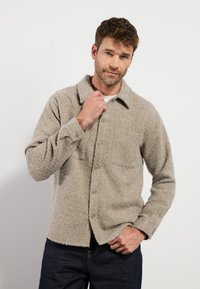 Veste chemise beige texturée avec un col, fermeture à boutons et une poche poitrine, assortie à un jean bleu foncé. Tissu doux et design décontracté.