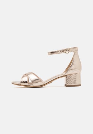 Metallic gold ankle strap sandal with a square toe, crisscross upper straps, and a textured surface, featuring a low block heel.