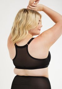 Musta langaton rintaliivi, jossa on läpinäkyvä selkä, säädettävät hihnat ja sileä pinta. Suunnittelu tarjoaa mukavan istuvuuden ja tuen.