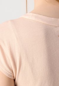 T-shirt à manches courtes, couleur pêche clair, avec un col ras du cou côtelé. Tissu en coton lisse avec une texture subtile et un petit détail imprimé au dos.