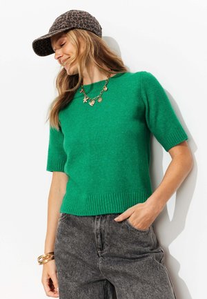 Femme portant un pull vert à manches courtes, un jean gris, une casquette à imprimé animalier, un collier porte-bonheur en or et des bracelets en or, souriante, la main dans la poche.